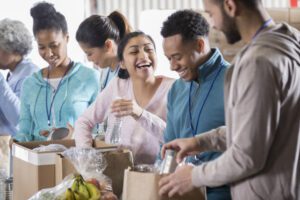 Cheerful young woman laughs along with her husband or boyfriend while volunteering in a community food bank. They are sorting through food donations. Volunteers are working in the background.